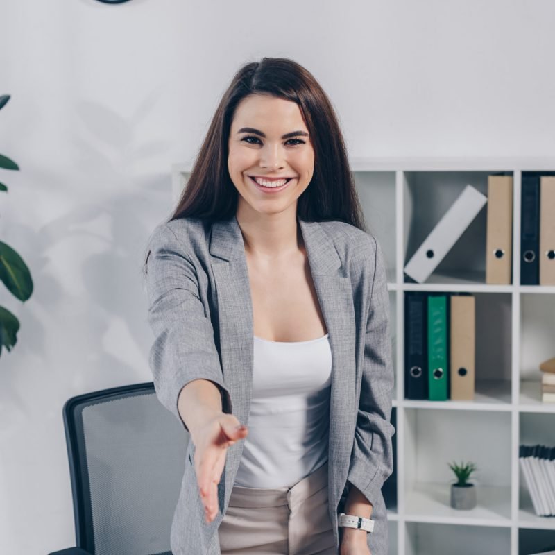 Recruiter with outstretched hand looking at camera and smiling at table in office